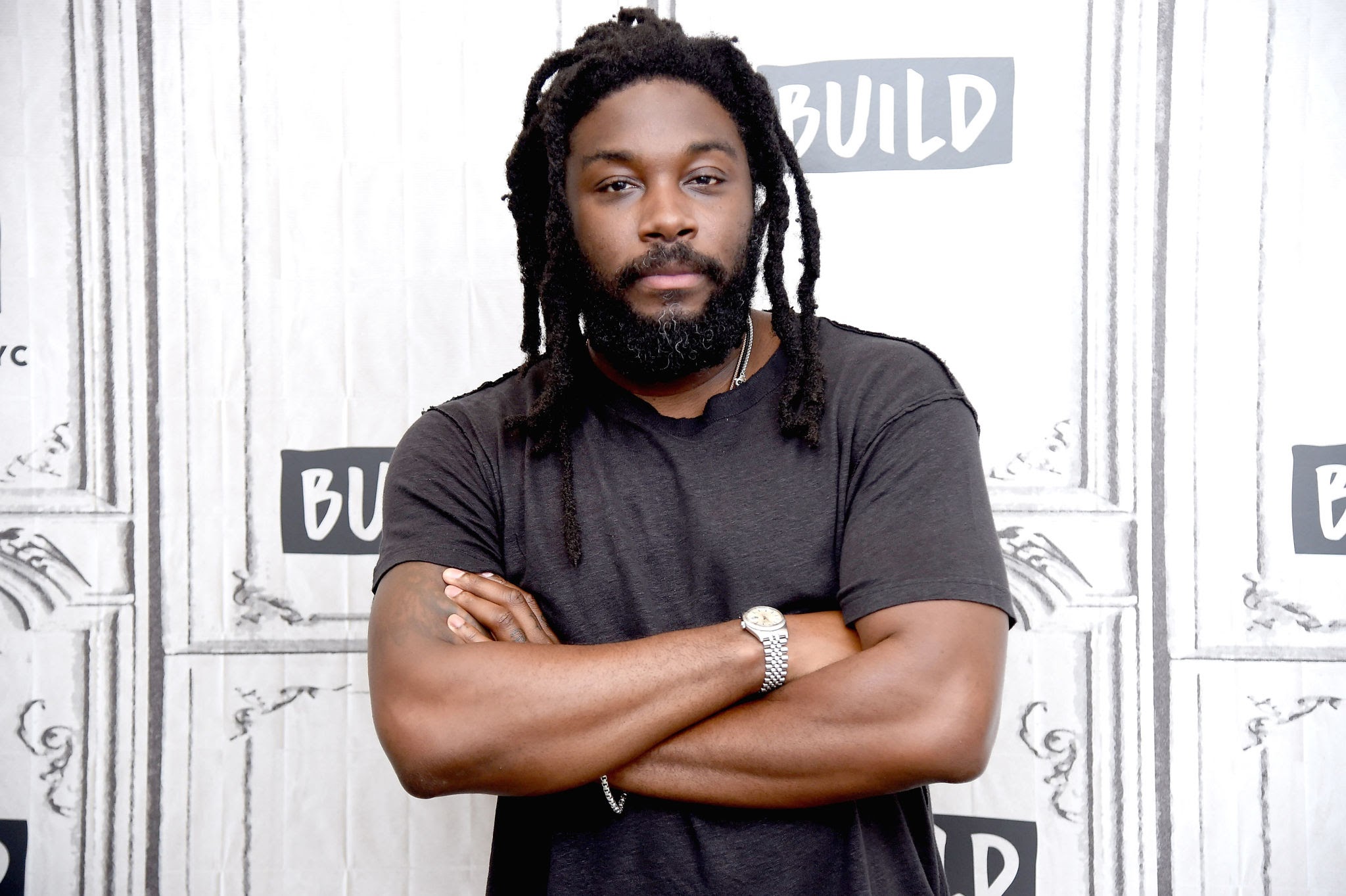 Author Jason Reynolds. He is a black man with a beard and locked hair looking straight at the camera. He is wearing a short sleeved black t-shirt, and has his arms crossed. 