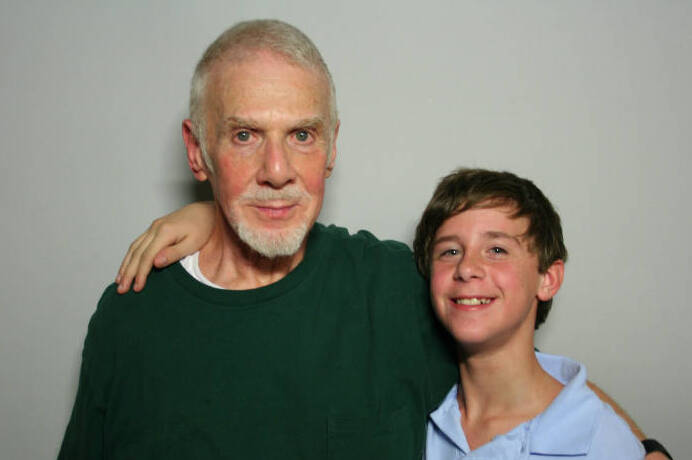 Elderly white man in green sweater posing for photo with younger white boy in light blue shirt against light background