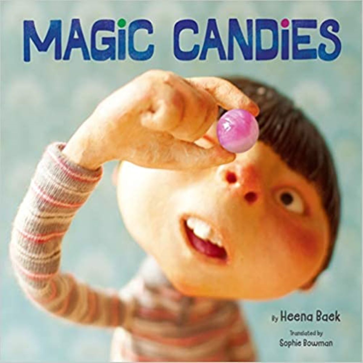 Book Cover of "Magic Candies" by Heena Baek; includes a boy with brown hair cut into a blunt bowl cut and wearing a long sleeved striped shirt. He is holding up a pink gumball to his eye and looking at it. 