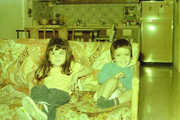 Picture of author as a child in Puerto Rico, with her younger brother. Picture shows two dark haired children sitting on a floral couch. 