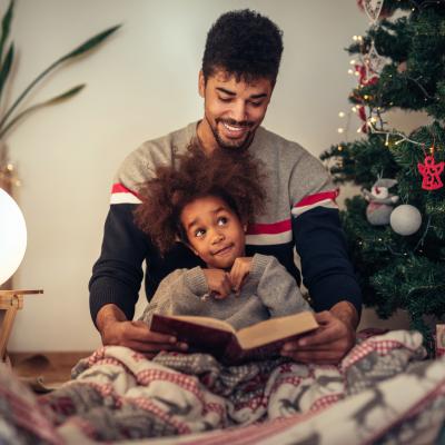 Father and son reading a book sitting by a Christmas tree. Son sits on father's lap, both are wearing pajamas.