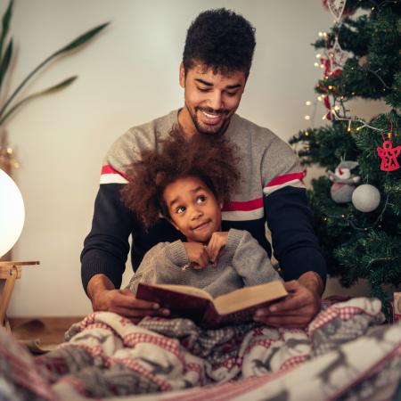 Father and son reading a book sitting by a Christmas tree. Son sits on father's lap, both are wearing pajamas.